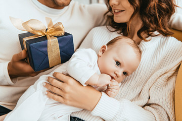 cropped view of husband presenting gift box to happy wife holding adorable baby