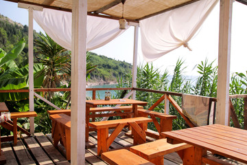 Tropical beach cafe bar at coastline by sea with outdoor view. Terrace balcony, relax place for eating and drinks of seaside resort with wooden fence, empty table and chairs and full sea panorama.