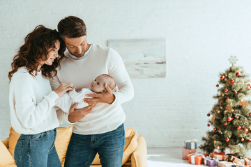 Happy parents standing near decorated Christmas tree and holding baby