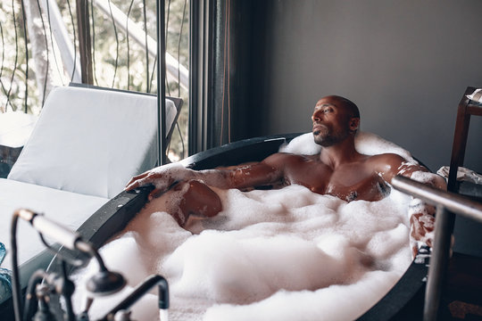 Brutal Arabic Man Having A Bath In His First Class Suit, The Guy Is Lying In The Black Stylish Tub In Thick Foam Near The Window; Luxury Concept.