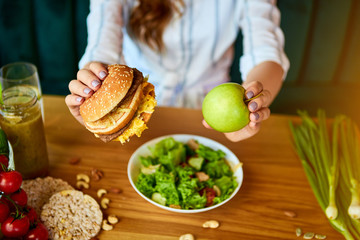 Beautiful young woman decides eating hamburger or apple in kitchen. Cheap junk food vs healthy diet