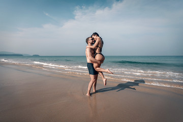 Strong man holds his girlfriend in his arms on the beautiful beach by the blue sea, love and tenderness.
