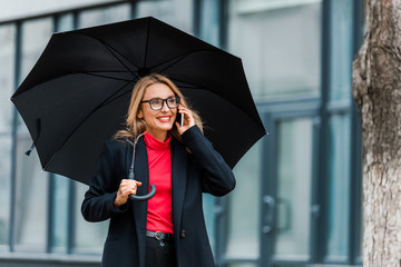 attractive and smiling businesswoman in black coat holding umbrella and talking on smartphone