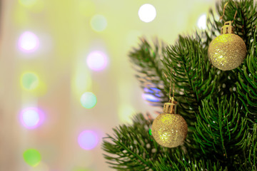 Two small cristmas-tree balls with golden glitter hang on needles of lush green Christmas tree branch. The background is blurred, bokeh
