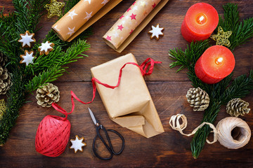 Wrapping Christmas gifts on a dark rustic wooden table surrounded by burning candles, fir branches, cinnamon stars and decoration, high angle top view from above