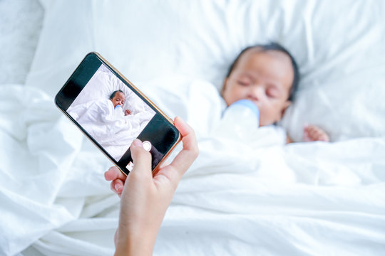 Mother hand hold mobile phone to take a photo of newborn baby during she is sleeping on white bed.