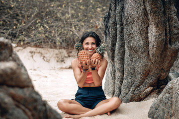Cheerful cute young woman in lotus pose at the beach holds two pineapples by her face; funny yoga concept