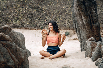 Beautiful girl in lotus pose looks at the pineapple, sitting at the beach.