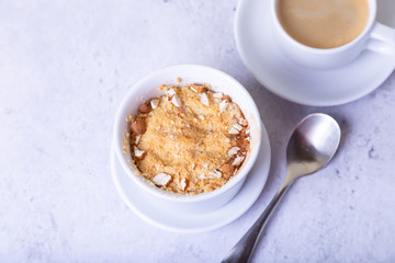Crumble with apples, cinnamon and almonds in a white ramekin. In the background is a cup of coffee. Close-up, selective focus.