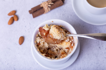 Crumble with apples, cinnamon and almonds in a white ramekin. In the background is a cup of coffee. Close-up, selective focus.
