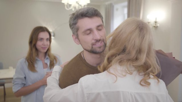 Handsome Caucasian man giving bouquet of violet roses to blond woman and hugging her. Guy showing annoyed facial expression, and smiling together with his young beautiful wife. Insincerity, hypocrisy