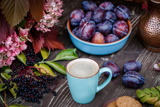 Fruits in a blue cup on a wooden table, plums, black elderberry, leaves of wild grapes, coffee, morning still life in the garden