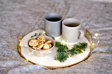On a marble tray with a gold rim, there are two gray cups of tea and a chocolate gingerbread cookie. A treat for Santa Claus. Christmas tree branch with garlands decorate the new year card. Copy space