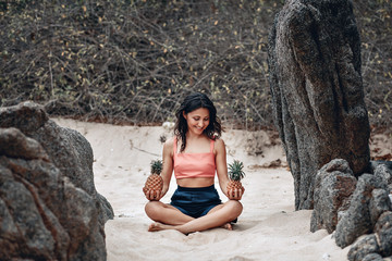 Smiling attractive lady in lotus pose at the beach looks at two pineapples in her hands; funny yoga concept