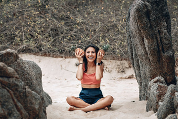Laughing girl in yoga pose at the beach holds two pineapples by her ears; funny sports concept