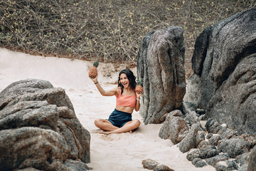 Laughing girl in lotus pose at the beach holds two pineapples; funny yoga concept