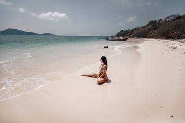 Fototapeta na wymiar Sexy lady in bikini sitting on clean sandy beach her leg stretched, wet body; style concept.