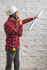 a civil engineer in a white helmet with documents in his hands talks on the phone at the white brick wall.