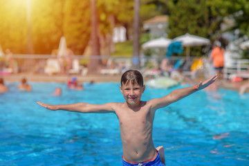 Obraz na płótnie Canvas Caucasian boy in blue swimming shorts jumping into swimming at resort in summer during his summer holidays. He is happy, his hands are open wide and he smiling towards camera.