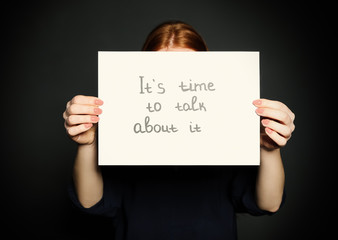 Young woman holding card with words IT'S TIME TO TALK ABOUT IT against dark background