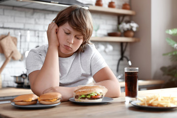 Emotional overweight boy at table with fast food in kitchen