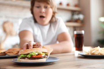 Emotional overweight boy reaching for burger at table in kitchen