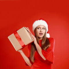 Emotional young woman in Santa hat with Christmas gift on red background