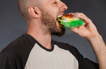 Environmental pollution and food industry. Close up portrait of a bald man with a beard, who enjoys biting into a hamburger filled with garbage and bags