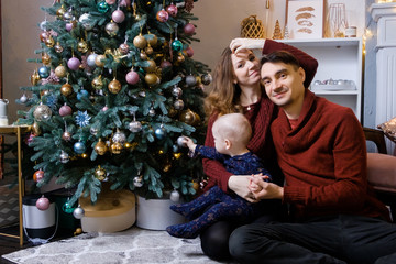 Beautiful young family sits on a carpet near the fireplace next to a large elegant Christmas tree