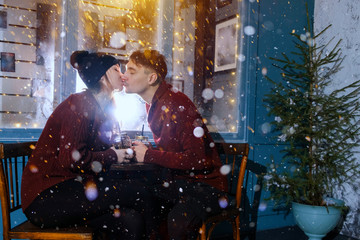 Young beautiful couple sits near an open-air cafe under snowfall. Loving couple kisses Christmas concept