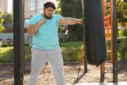 Young Overweight Man Kicking Heavy Bag On Sports Ground