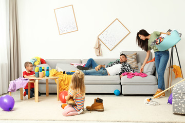 Exhausted mother trying to clean up mess made by children and lazy father in room
