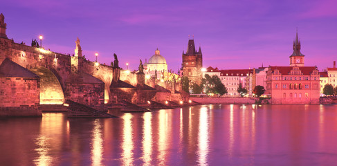 Fototapeta na wymiar Prague on sunset, illuminated Charles bridge, Bridge Tower and old buildings on the riverside under purple sky with golden reflections in Vltava river, panoramic image