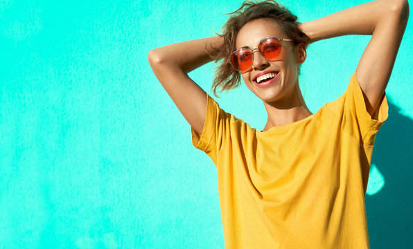 Fashionable cheerful young woman posing on blue background, wearing yellow t-short and trendy red eyeglasses.