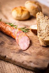 A rustic composition. Sliced, smoked pork sausage, fresh herbs and bread on a wood cutting board and jute fabric.