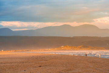 Fototapeta na wymiar A streak of golden evening light falling on the Plettenberg Bay beach at sunset, with mountains in the distance and people walking on the beach. Garden Route, Western Cape, South Africa