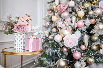 New Year's holiday bouquet in a vase, a gently pink gift present box on the bedside glass table against the background of a decorated Christmas tree in soft pink colors. New Year and Christmas concept