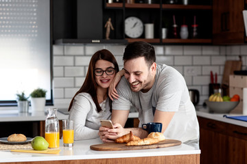 Couple in the kitchen eat breakfast with orange juice and pastry at table with mobile phone on hand smile