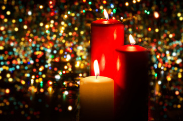 Three Christmas candles on a dark background of blurred colored garland lights. Composition of festive mood. Coming new year. Interior decoration and romantic atmosphere. Copy space