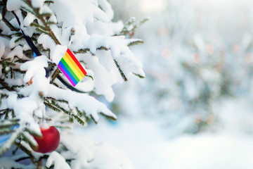 Christmas LGBT. Xmas tree covered with snow, decorations and rainbow flag. Snowy forest background in winter. Christmas greeting card. - 299389785
