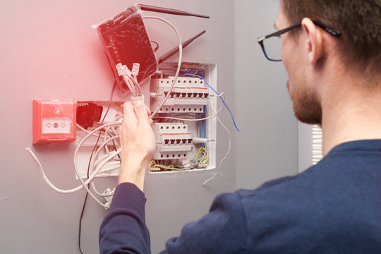 A man with glasses opposite the circuit breakers with wires and a router. Inexperienced electrical engineer in front of the electrical panel in the house. - Powered by Adobe
