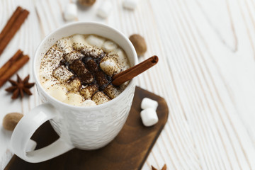 White mug with hot chocolate and marshmallows with cinnamon stick on a wooden table. Hot winter drink, top view.