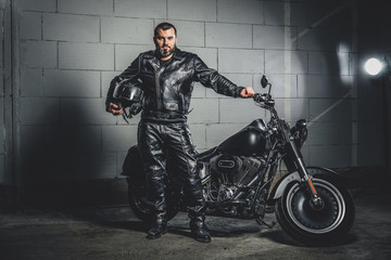 Brutal bearded biker in leather suit is standing next to his bike while holding a helmet.