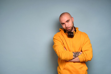 A guy with a beard in a yellow sweatshirt is standing on a blue background with headphones on his neck, looking at the camera.