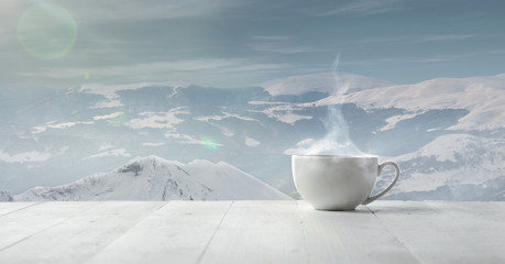 Single tea or coffee cup and landscape of mountains on background. Cup of hot drink with snowly look and cloudly sky in front of it. Warm in winter day, holidays, travel, New Year and Christmas time.