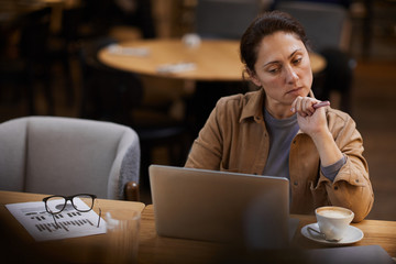 Serious businesswoman concentrating on her online work she working on laptop at the table with financial graphs and coffee cup in cafe