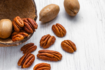 Pecans peeled and in shell lying in half the coconut shell and on a white wooden surface, side view from above