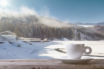 Obraz na płótnie Canvas Single tea or coffee cup and landscape of mountains on background. Cup of hot drink with snowly look and cloudly sky in front of it. Warm in winter day, holidays, travel, New Year and Christmas time.