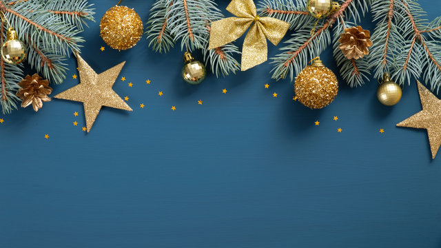 Christmas frame top border made of fir tree branches, golden decorative stars, balls over blue background. Flat lay, top view. Xmas banner mockup with copy space