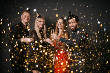 Obraz na płótnie Canvas Two attractive guys and two beautiful girls are clinking with champagne glasses and celebrating new year. On friends from above the festive confetti falling. Shooting on isolated black background.